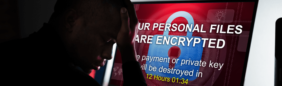 man holding his head, with a computer with a ransomware alert on the screen behind him